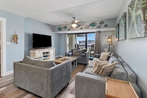 A living room with a flat-screen TV, grey sofas, a wooden coffee table, and a ceiling fan. - The Club at Mexico Beach, 3BD/2BA, panoramic gulf views, steps to the beach, community pool! (Mexico Beach)