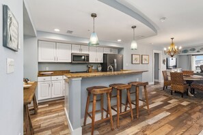 A modern kitchen with white cabinets, a central island with stools, a dining area with wicker chairs, and a chandelier. - The Club at Mexico Beach, 3BD/2BA, panoramic gulf views, steps to the beach, community pool! (Mexico Beach)