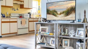 Una sala de estar con una televisión que muestra una escena de playa, un estante de madera con diversos objetos decorativos, y una cocina al fondo.