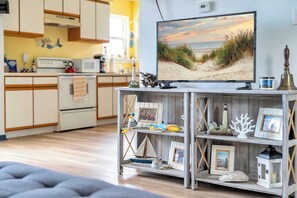 A living room with a television displaying a beach scene, a wooden shelf with various decorative items, and a kitchen in the background.