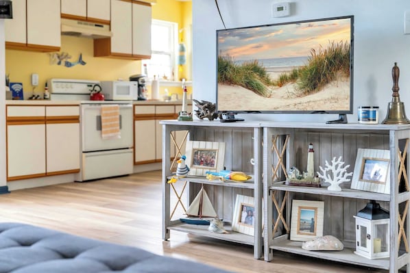 A living room with a television displaying a beach scene, a wooden shelf with various decorative items, and a kitchen in the background. - 2 Bedroom Bay Escape (Middle Township)