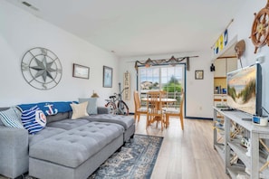 A living room with a grey sectional sofa, a dining area with wooden chairs, a bicycle, and a TV on a wooden stand.