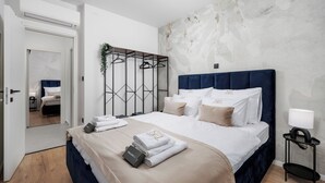 Una habitación de hotel moderna con una cama grande, un armario, una mesita de noche con lámpara y vistas a otra habitación a través de una puerta abierta.