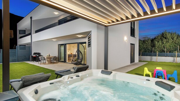Un área moderna de hidromasaje al aire libre con un espacio de estar cubierto, un patio con una parrilla y vistas a la casa.