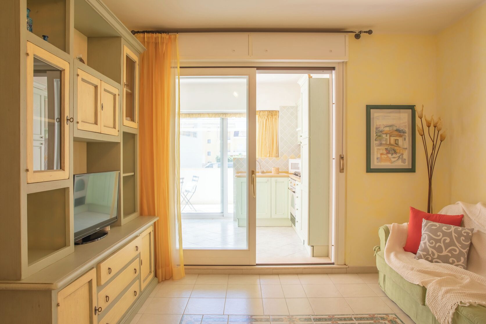 Een woonkamer met een televisie, een bank, een schuifdeur naar een balkon en een zichtbare keuken door de deur.
