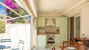 Een kleine keuken met een fornuis, magnetron en kastjes, een eettafel met stoelen, en uitzicht op een balkon met bloemen.