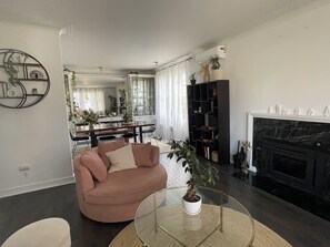 Un salon avec un canapĂ©, une table basse ronde en verre agrĂ©mentĂ©e dâune plante, une cheminĂ©e et, au fond, un coin repas.
