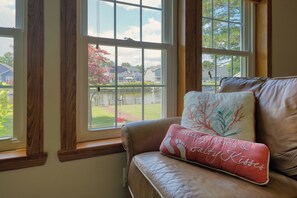 A leather sofa with a cushion that reads 'Sandy Toes & Happy Kisses' and a window with a view of a residential area. - The Hoagie House - Family-Friendly Canal Home with Private Dock (Frankford)