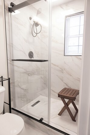 A glass-enclosed shower with a marble wall, a wooden stool, and a window with blinds.