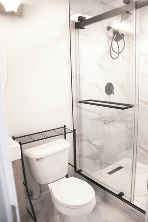 Ein modernes Badezimmer mit einer gläsernen Duschkabine, einer weißen Toilette und einem schwarzen Metallregal.