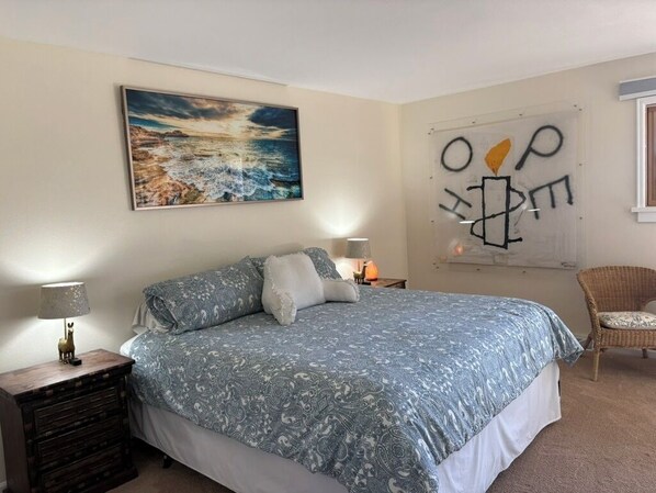 Ein Schlafzimmer mit Bett, Nachttisch, Lampe, Sessel, einem Gemälde und einer Wand mit abstrakter Kunst.