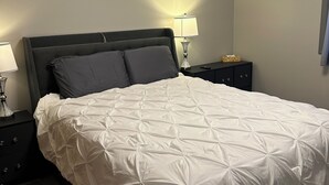 Een net opgemaakt bed met een witte deken, twee bedlampen, een klein nachtkastje en een nachtkastje in donkere tinten.