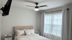 Un dormitorio con cama, ventilador de techo, mesita de noche, ventana con cortinas e iluminación empotrada en la pared.