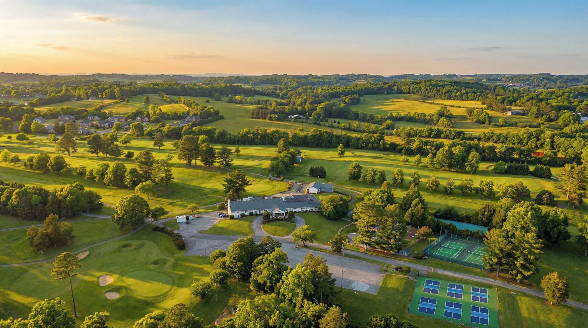 An aerial view of a golf course with a clubhouse, tennis courts, and surrounding greenery.