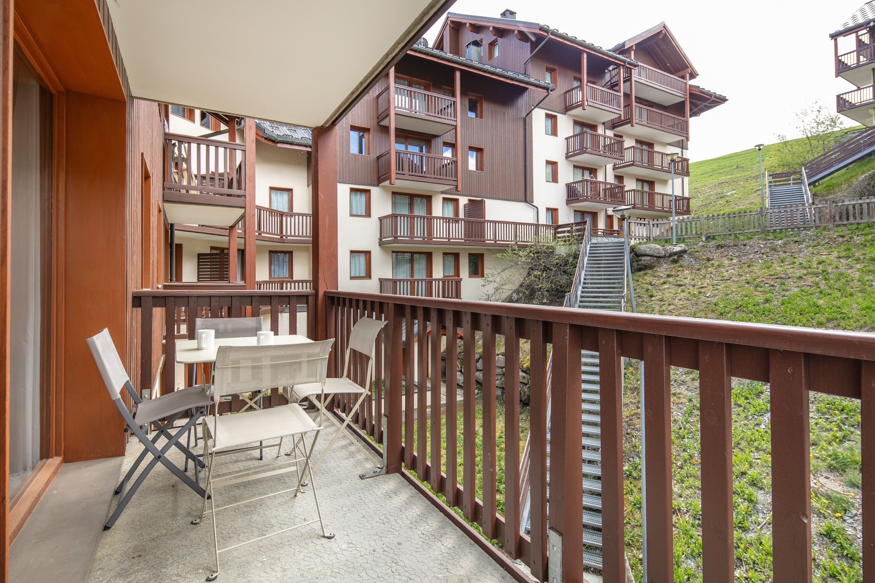 A multi-story building with balconies, a terrace with chairs and a table, and a staircase leading to a hill.