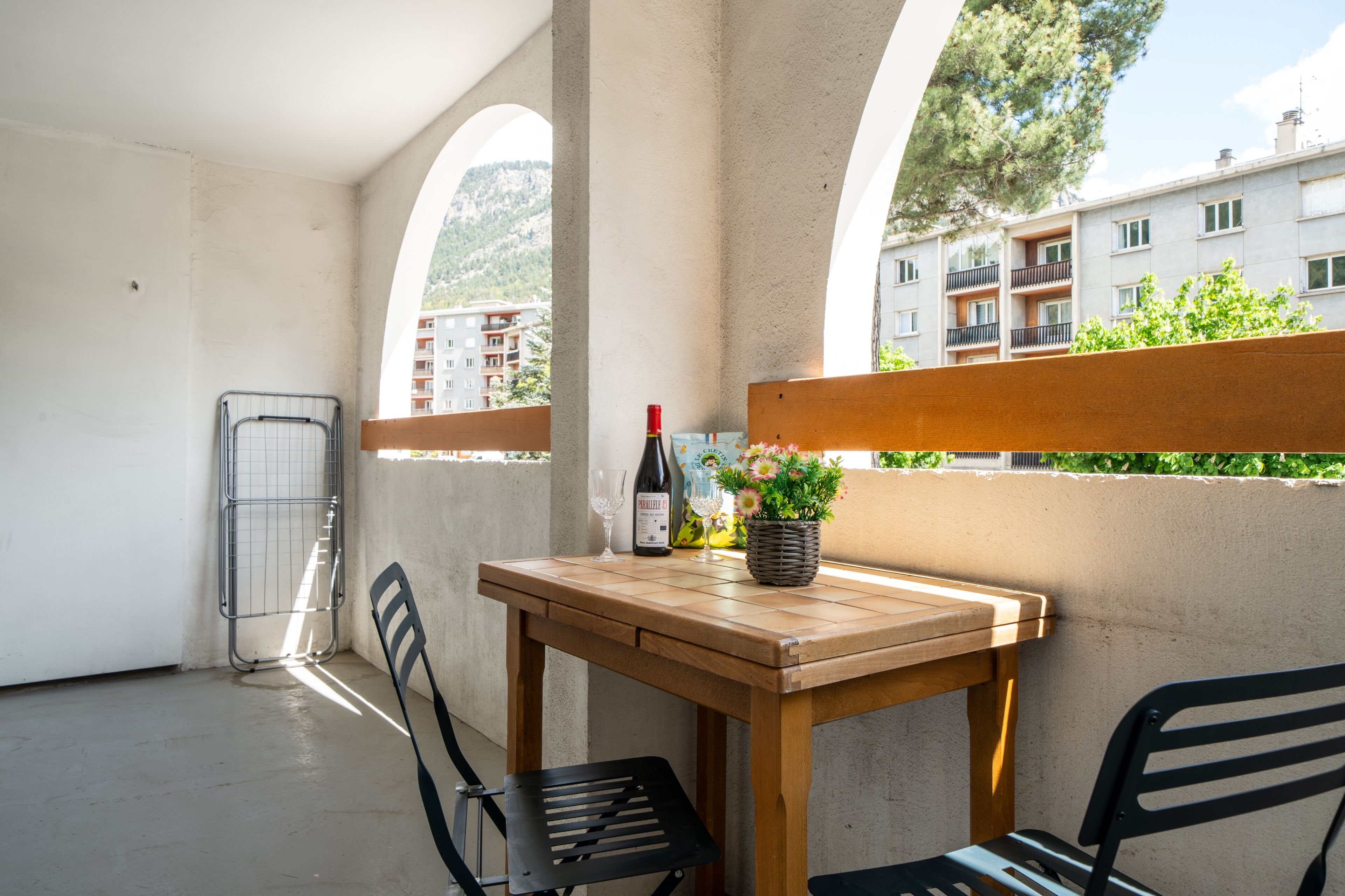 Een balkon met een houten tafel, twee stoelen, een fles wijn en een bloevas.