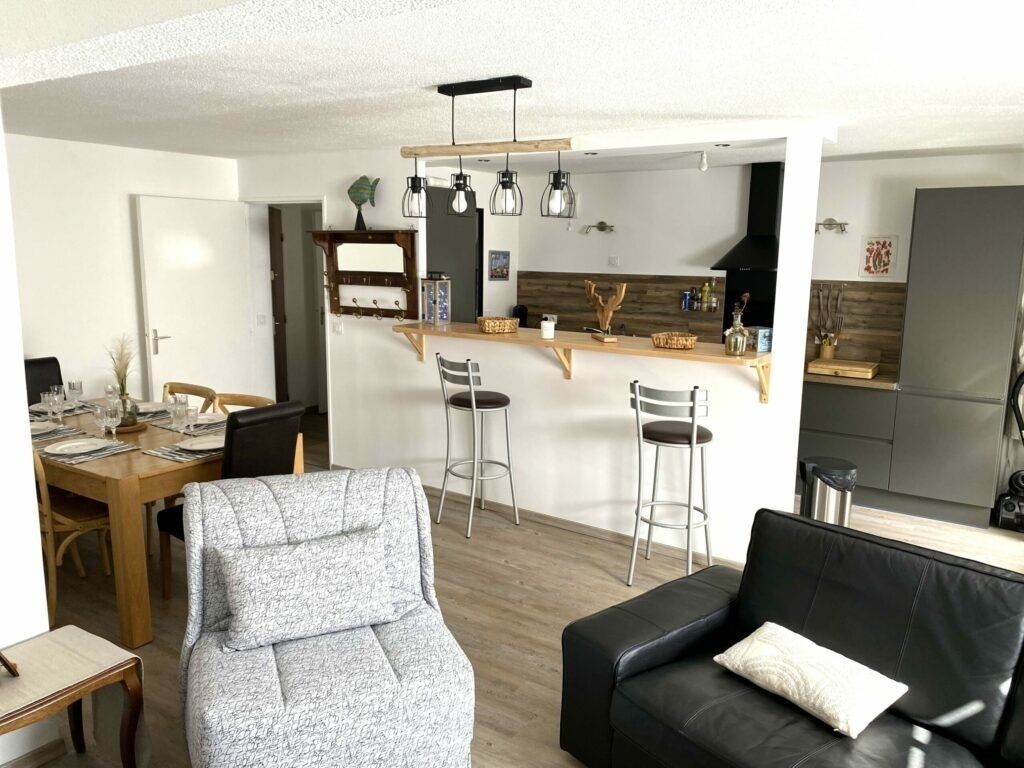 Ein modernes Wohnzimmer mit einer schwarzen Ledersofa, einem grauen Sessel und einem Esstisch mit Stühlen. Im Hintergrund ist eine Küchenzeile mit einer Holz Arbeitsplatte und Barhocker zu sehen.
