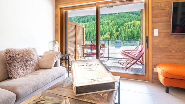 Ein Zimmer mit einer Couch, einem Buch, Holzboden, einer Schiebetür aus Glas und Blick auf einen Wald.