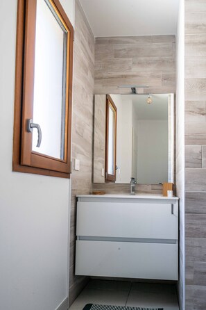 Ein Badezimmer mit einem raumhohen Holzfenster, einem weißen Waschbecken mit Spiegel darüber und einer hell gefliesten Wand.