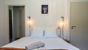 Un llit amb llençols blancs, dues làmpades de nit, una tovallola plegada i un quadre a la paret.