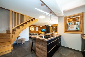 A modern kitchen with a wooden staircase, a central island, pendant lighting, and a television in the background.