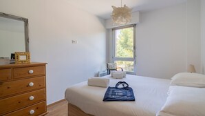 Ein Schlafzimmer mit einem Holzkasten, einem Bett mit weißer Bettwäsche, einer Pendelleuchte und einem Fenster mit Blick auf Bäume.