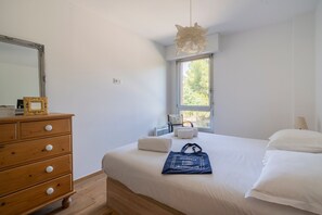 A bedroom with a wooden dresser, a bed with white linens, a hanging light fixture, and a window with a view of trees. - Match Point - À moins d'1 km des plages (Marseille)