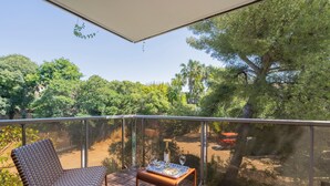 Ein Balkon mit einem Korbstuhl, einem kleinen Tisch mit Flasche und Gläsern sowie Blick auf Bäume und ein Gebäude.