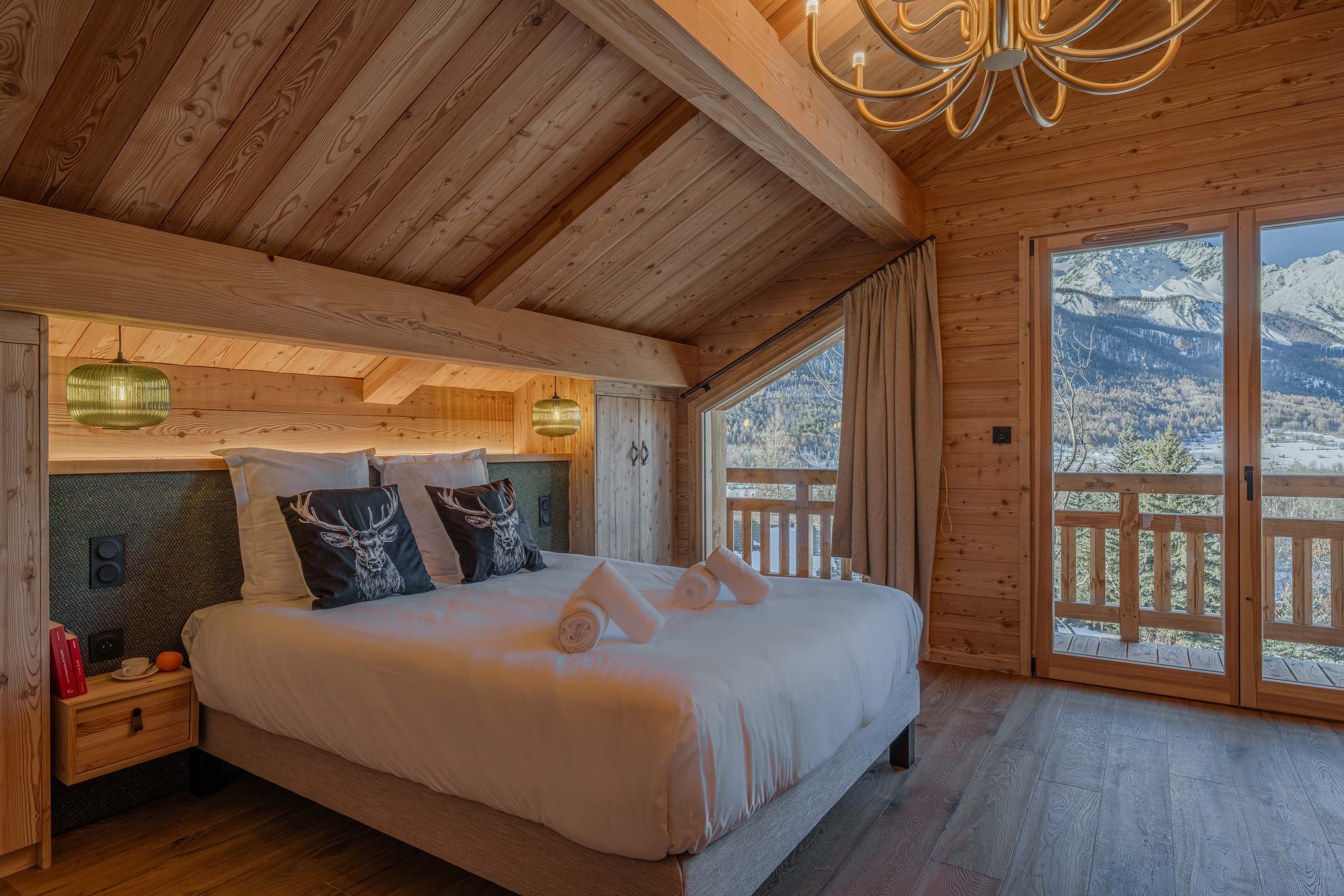 A wooden cabin bedroom with a bed, bedside tables, and a view of snow-capped mountains through large windows.