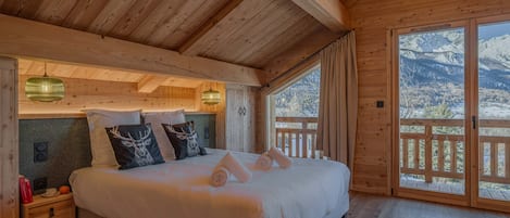 A wooden cabin bedroom with a bed, bedside tables, and a view of snow-capped mountains through large windows.