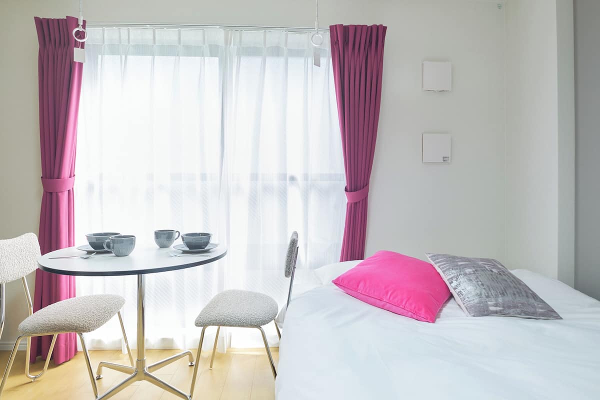Quarto com cama, mesa redonda com cadeiras, janela com cortinas leves e interruptor de luz na parede.