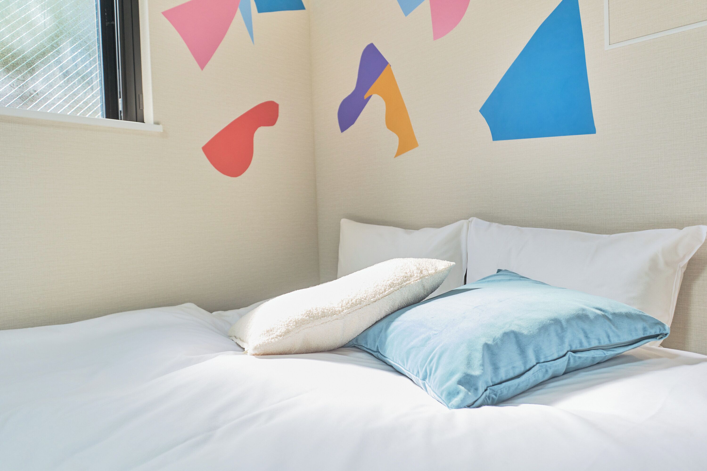 A bed with white and blue pillows, a window with blinds, and a wall with abstract shapes.
