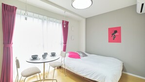 A room with a bed, a round table with chairs, a window with curtains, and a wall-mounted light fixture.