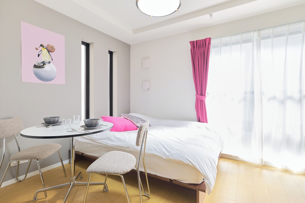Un dormitorio moderno con cama, una mesa redonda de comedor con sillas, una ventana con cortinas translúcidas y una obra de arte colgada en la pared.