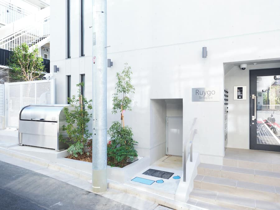 A modern building entrance with a sign 'Ruygo,' a metal mailbox, and a small garden area.