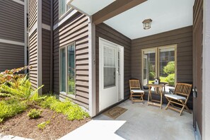 A covered patio with a door, two wooden chairs, a small table, and a doormat. - Newly Renovated 1st Floor Condo | Walk to Main St. (Blowing Rock)