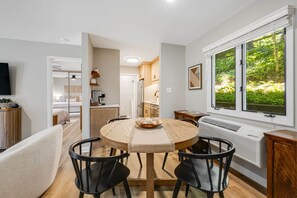 A dining area with a round table, four chairs, a central decorative bowl, a window with a view of greenery, and a built-in air conditioning unit.