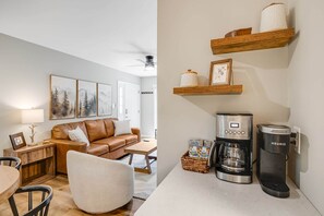 A living room with a brown leather sofa, a white armchair, a wooden coffee table, and a wall-mounted shelf with a coffee maker and decorative items. - Newly Renovated 1st Floor Condo | Walk to Main St. (Blowing Rock)