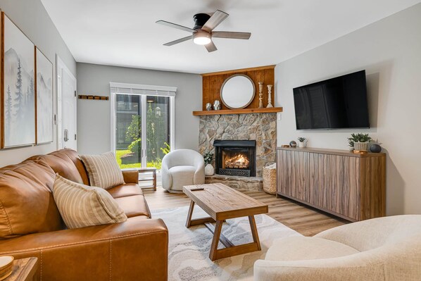 A living room with a leather sofa, a white armchair, a wooden coffee table, a stone fireplace, a TV mounted on the wall, and a ceiling fan.