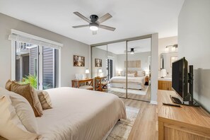 Quarto com cama, televisão, ventoinha no teto, escrivaninha e espelho.