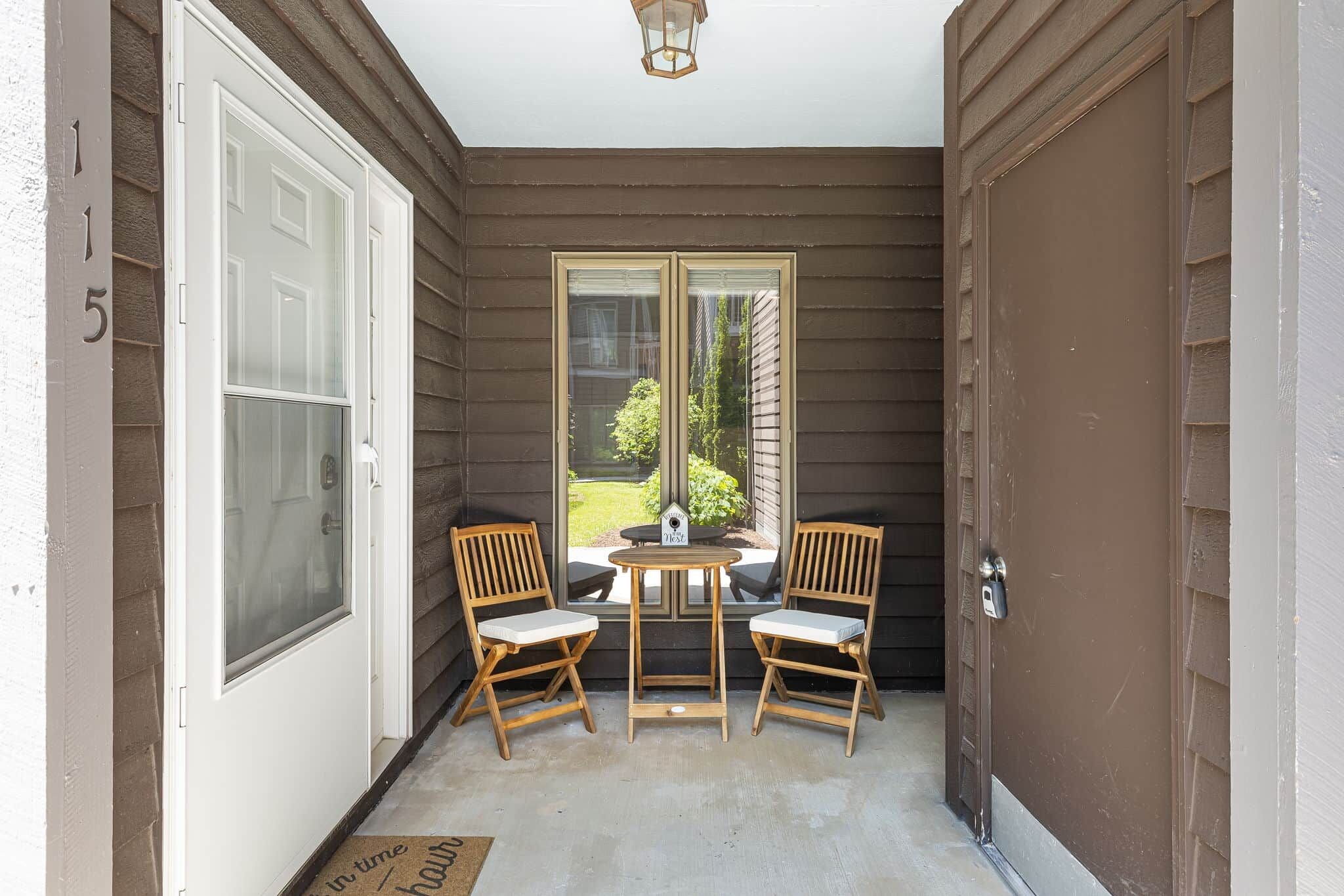 A covered porch with a door, two chairs, a small table, and a view of the outside.
