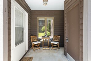 A covered porch with a door, two chairs, a small table, and a view of the outside.