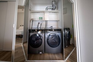 A compact laundry room with two front-loading washing machines, a small shelf with a detergent packet, and a ventilation system above. - Condo in the heart of Westboro (Ottawa)