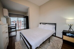 A bedroom with a bed, a desk, and a chair, a window with a view of buildings, and a lamp on a nightstand. - Spacious Apartment near University of Ottawa (Ottawa)