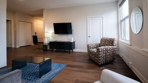 Ein Wohnzimmer mit Flachbildfernseher, einem gemusterten Sessel, einer Couch, einem Glastisch und einem blauen Teppich.
