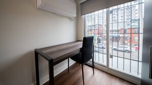 Ein Zimmer mit einem Holztisch, einem schwarzen Stuhl, einem großen Fenster mit Blick auf Gebäude und eine Straße sowie einer weißen Klimaanlage.