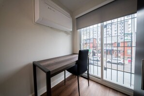 Ein Zimmer mit einem Holztisch, einem schwarzen Stuhl, einem großen Fenster mit Blick auf Gebäude und eine Straße sowie einer weißen Klimaanlage.