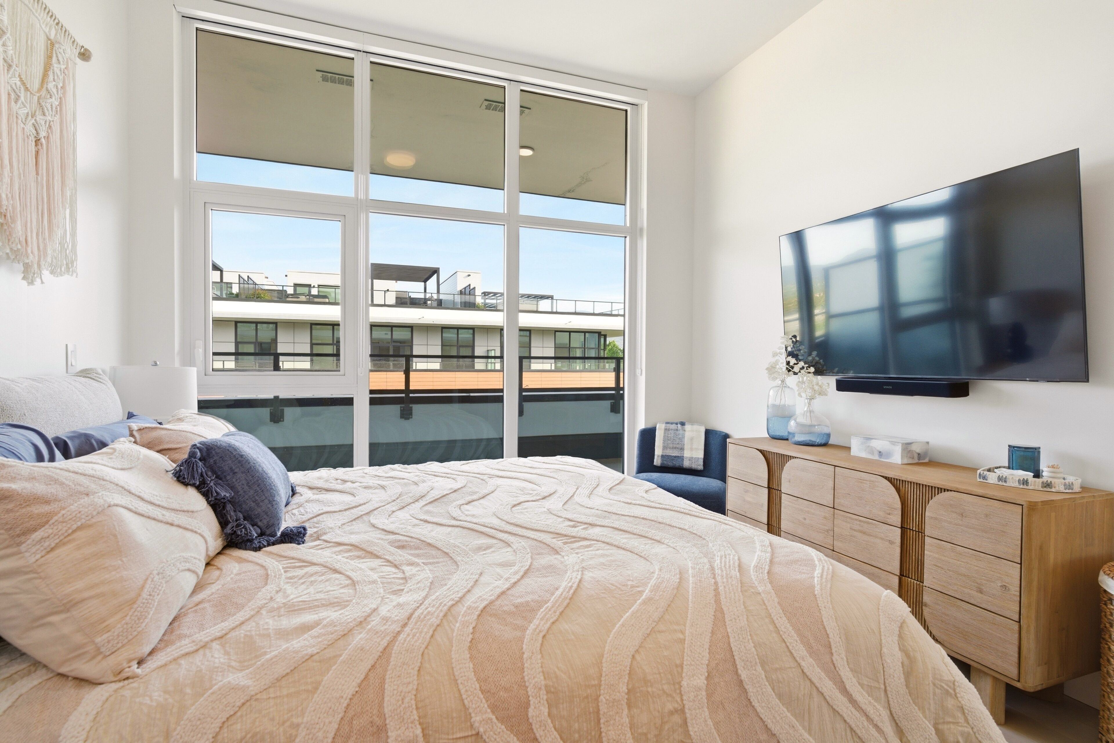 Une chambre avec un grand lit, une télévision, une commode en bois et un balcon offrant une vue sur les immeubles.