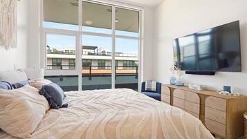 Une chambre avec un grand lit, une télévision, une commode en bois et un balcon offrant une vue sur les immeubles.