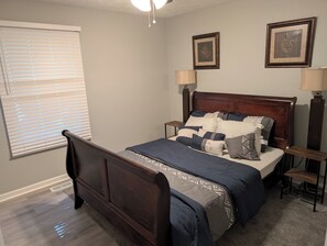 A bedroom with a wooden bed, two wall-mounted lamps, a window with blinds, and two framed pictures on the wall. - Bring the crew to this comfortable 3/2 home minutes from everything in Anniston (Anniston)